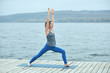 © sandsun - Beautiful young woman practices yoga asana Virabhadrasana 1 - warrior pose 1 on the wooden deck near the lake