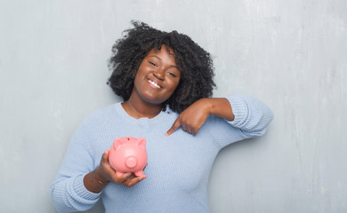 Sticker - Young african american woman over grey grunge wall holding piggy bank with surprise face pointing finger to himself