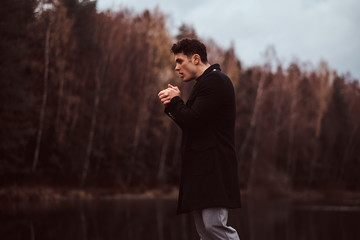 A handsome young man warms hands near a lake in the autumn forest.