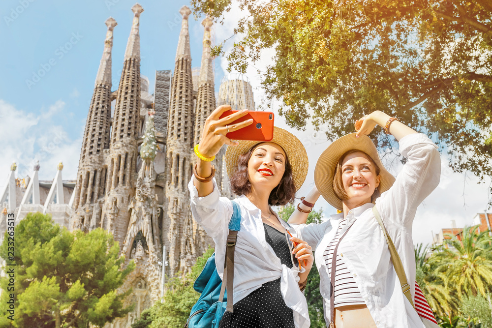 Obraz na płótnie BARCELONA, SPAIN - 11 JULY 2018: Young girls friends making selfie photo on her smartphone in front of the famous Sagrada Familia catholic cathedral. Travel in Barcelona concept w salonie