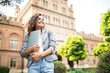 © F8  \ Suport Ukraine - Smiling brunette student walking outdoor with laptop and backpack and looking in smartphone in sunlight on background of university