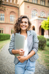 Portrait of young beautiful woman student walking in the park using mobile phone holding laptop computer.