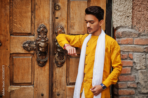 Fotografie, Obraz Indian stylish man in yellow traditional clothes with white scarf posed outdoor against old gates