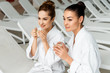 © LIGHTFIELD STUDIOS - beautiful young women in bathrobes holding cups with herbal tea in spa
