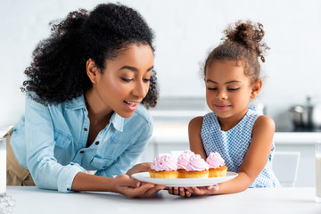 Wall Mural - african american mother and daughter looking at homemade cupcakes in kitchen