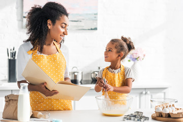 african american mother holding cookbook and daughter preparing dough in kitchen, looking at each ot
