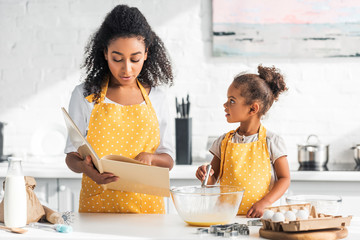 african american mother reading cookbook and daughter whisking eggs for dough in kitchen