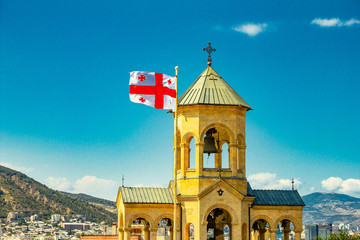 Beautiful view to Georgian Flag on the background chapel on site to Sameba Cathedral Tsminda in Tbilisi Holy Trinity . Biggest church Orthodox in Caucasus Georgia region in a sanny day.