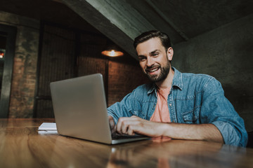 I love my job. Portrait of outgoing bearded male employer with attractive smile having work with laptop while locating at desk. He wearing casual clothes