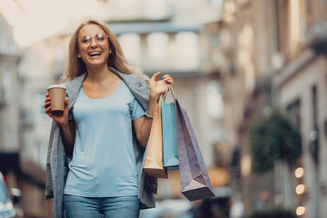 Waist up portrait of beautiful middle-aged lady in glasses holding cup of coffee and shopping bags. She is looking at camera and laughing