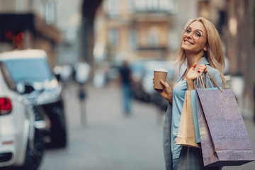 Waist up portrait of charming middle-aged lady holding cup of coffee and shopping bags. She is looking at camera and smiling