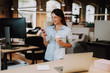 © Yakobchuk Olena - Waist up portrait of charming woman using smartphone while holding cup of coffee. She is standing near desk with laptop and smiling