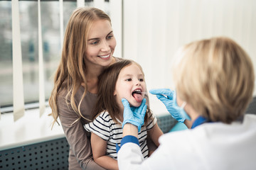 Wall Mural - Concept of professional consultation and examination. Waist up portrait of little girl with opening mouth being examined by pediatrician woman in medical office