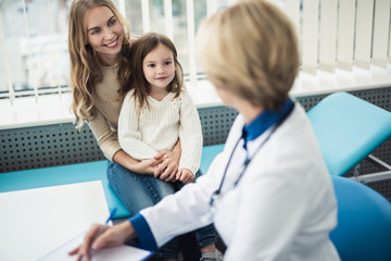 Wall Mural - Concept of professional consultation in healthcare system. Portrait of smiling little girl with her mother being consulting by pediatrician woman in medical office