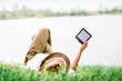 Girl reading e-book lying on the grass by the lake.