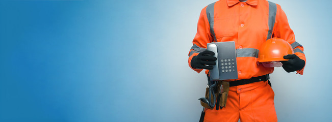 Wall Mural - Builder worker is holding in hand a handset phone and hard hat isolated on blue background with copy space. Call to a repair man concept. Contact us.