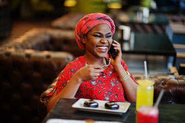 Wall Mural - Stylish african woman in red shirt and hat posed indoor cafe, eat chocolate dessert cakes and speaking mobile phone.