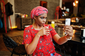 Wall Mural - Stylish african woman in red shirt and hat posed indoor cafe, drinking strawberry lemonade and making selfie on phone.