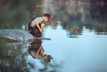 Caveman Boy Sitting On The Rock And Looking At Him Self In The Water Reflection In Lake. Evolution Survival Concept. Creative Art Fantasy Photo