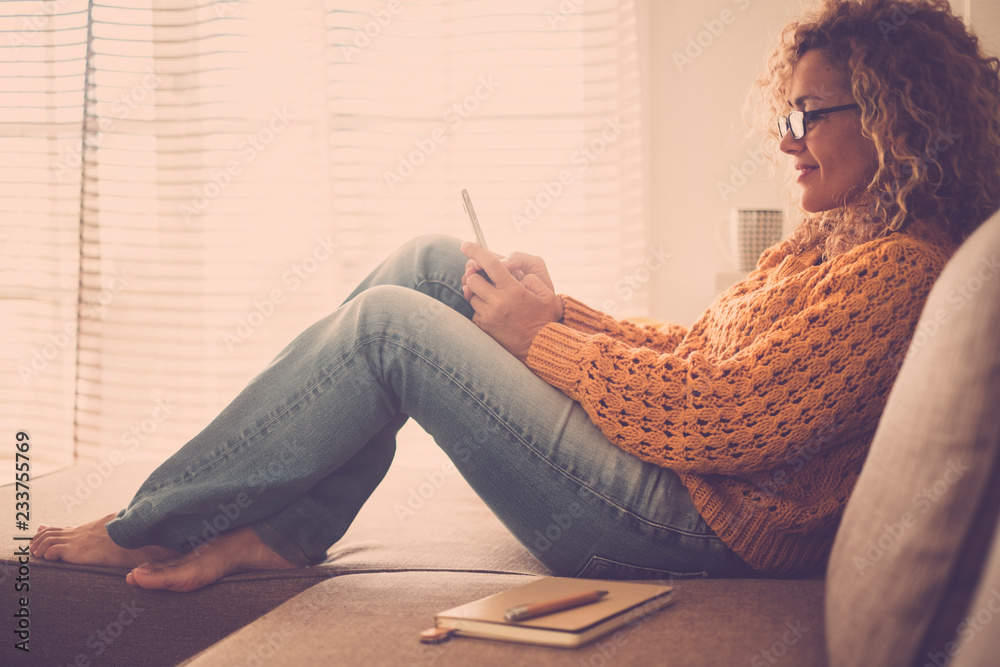 beautiful middle age caucasian woman at home lay on the sofa usingmobile phone internet technologhy - paper note and pen on the sofa - barefoot enjoying the living room - vintage warm colors - obrazy, fototapety, plakaty 