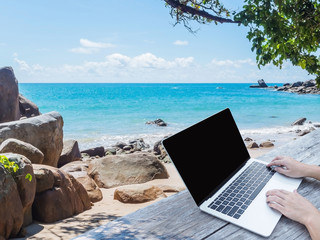 hand typing laptop with blank screen on wooden table over summer beach background.