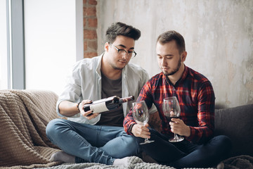 Wall Mural - Two sexy caucasian guys toasting with red wine glasses, engaging in home romantic party, celebrating their first month anniversary love.