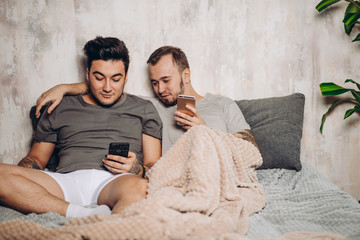 Wall Mural - Two contented homosexual male people lying together in bed and using smartphone to make video call to friends