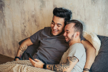 Wall Mural - Two contented homosexual male people lying together in bed and using smartphone to make video call to friends