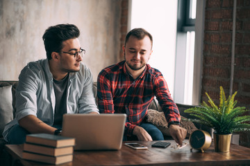 Wall Mural - Romantic partners in love and business. Caucasian amused gay couple looking at laptop screen while resting in living room with loft interior. Dominant man wearing spectacles and and using laptop