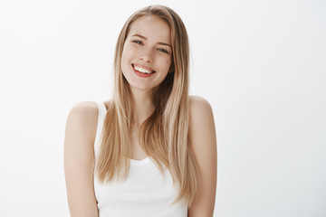 Wall Mural - Friendly-looking sociable young woman with blond hair and perfect skin smiling broadly with white teeth at camera, tilting head gazing happily, posing over gray background delighted and self-aware
