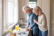 © fizkes - Smiling aged wife hug man from behind watching him preparing healthy food, loving senior woman embrace husband cooking breakfast slicing fruit, romantic couple spend morning at home together