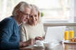 Aged couple busy look at laptop screen while having delicious breakfast at home kitchen, excited senior man and woman use computer during morning routine, elderly wife show something to husband at pc