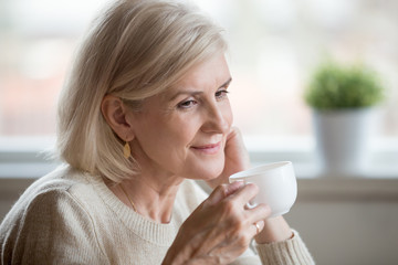 Close up of beautiful aged woman drinking tea thinking about pleasant moments, thoughtful senior female enjoying coffee remembering or dreaming about something, having calm morning at home