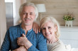 © fizkes - Portrait of happy senior couple posing for family picture at home, smiling elderly wife leaning on caring husband holding hands looking at camera during photoshoot, aged man and woman shoot embracing