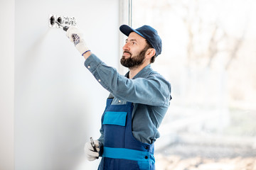 Electrician in uniform mounting electric sockets on the white wall indoors