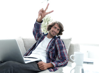 Wall Mural - happy guy with laptop sitting on sofa and showing his hand a winning gesture