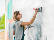Beautiful Young Blonde Girl Making Graffiti Of Big Eye With Aerosol Spray On Urban Street Wall. Creative Art. Talented Student In Denim Overalls Drawing Picture