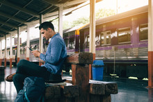 Asian Man Sitting On Bench And Using Smartphone With Waiting Train.