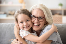 Happy Old Grandmother Hugging Little Grandchild Girl Looking At Camera, Smiling Mature Mother Or Senior Grandma Granny Laughing Embracing Adopted Kid Granddaughter Sitting On Couch, Headshot Portrait
