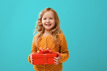 happy little child girl in a warm mustard knitted sweater holding a red box with a gift on a cyan ba