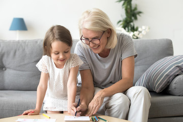 Wall Mural - Smiling grandmother and little kid granddaughter drawing on paper with colored pencils together, caring granny teaching grandchild having fun playing at home, grandma and creative child activity