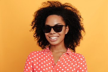 Wall Mural - Woman portrait. Afro American girl in sun glasses is looking at camera and smiling, on a yellow background