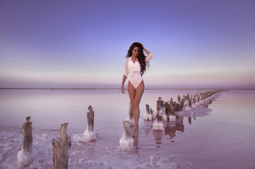 Wall Mural - Sexy woman in white bikini posing on salty lake with pink water and the beach from salt. Nature miracle. Beautiful tanned girl with long curly healthy hair. Wellness SPA treatment concept.