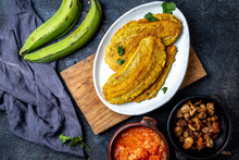 COLOMBIAN CARIBBEAN CENTRAL AMERICAN FOOD. Patacon Or Toston, Fried And Flattened Whole Green Plantain Banana On White Plate With Tomato Sauce And Chicharron Black Background, Top View