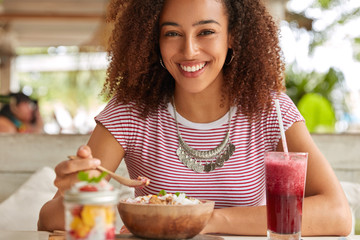 Pretty young female model holds wooden spoon, tastes delicious exotic salad with fruit, being in high spirit, drink fresh cocktail, looks happily at camera, poses against blurred cafe background