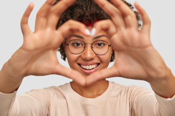 Photo of attractive young woman makes heart shape gesture over face, smiles broadly, demonstrates her love to boyfriend, wears round spectacles, isolated over white background, gestures indoor