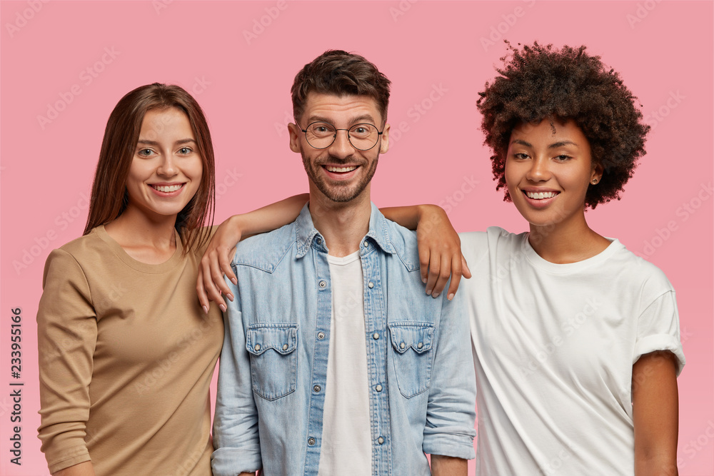 Horizontal shot of three mixed race teenagers spend time together, pose for common photo against pink background. Satisfied guy in eyewear and denim shirt stands between two cheerful women indoor - obrazy, fototapety, plakaty 
