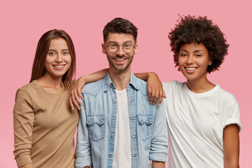 Wall Mural - Horizontal shot of three mixed race teenagers spend time together, pose for common photo against pink background. Satisfied guy in eyewear and denim shirt stands between two cheerful women indoor