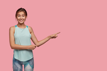 Wall Mural - Positive Caucasian woman with cheerful expression, dressed in casual vest and leggings, points aside with both index fingers, stands against pink background, has healthy lifestyle, likes sport