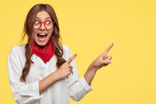 Photo Of Attractive Woman Has Dark Hair Slightly Combed In Plaits, Dressed In White Shirt And Stylish Bandana Around Neck, Points With Both Fore Fingers Against Yellow Background For Advertisement